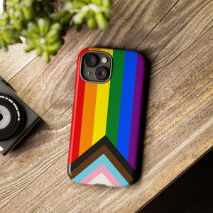 Puede incluir: Una funda para smartphone con un diseño de bandera arcoíris, con rayas verticales rojas, naranjas, amarillas, verdes, azules y moradas. La funda también incluye la bandera del Progreso del Orgullo con chevrones negros, marrones, celeste, rosa y blancos. La funda está sobre una superficie de madera.