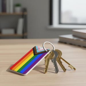 May include: A rectangular keychain featuring the rainbow flag and the progress pride flag. The keychain is attached to a silver keyring with several keys. The design includes red, orange, yellow, green, blue, and purple stripes.