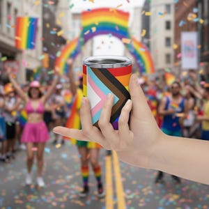 Puede incluir: Un vaso de acero inoxidable con el diseño de la bandera del Orgullo Progresista. El vaso se sostiene frente a una multitud que celebra un desfile del Orgullo, con banderas arcoíris y confeti en el fondo.