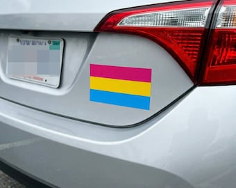 May include: A rectangular car sticker displaying the pansexual pride flag, with horizontal stripes of pink, yellow, and blue. The sticker is attached to the rear of a silver car, near the rear light.