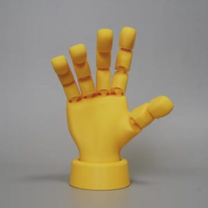 Può includere: Un modello di mano articolata gialla, con ogni dito e il pollice segmentati per il movimento. La mano è montata su una base circolare e le dita sono leggermente curve. Lo sfondo è grigio uniforme.