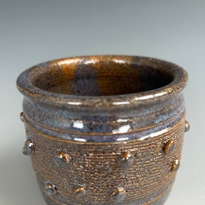 May include: A small, brown and blue ceramic pot with a textured surface and raised dots around the rim.
