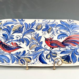 Puede incluir: Una fuente ovalada de cerámica con base blanca, con un diseño pintado a mano de hojas azules y pájaros rojos. Los pájaros tienen plumas de cola rojas y están posados entre el follaje. La fuente está soportada por dos soportes metálicos dorados.
