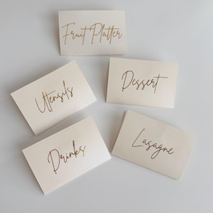 May include: Five rectangular off-white cards with gold foil lettering. The cards feature the text Fruit Platter, Utensils, Dessert, Drinks, and Lasagne in a cursive script font. Each card is folded and arranged in a scattered layout.