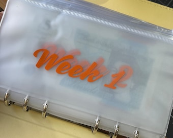 May include: A clear plastic zippered pouch with a label that says "Week 1" in orange lettering. The pouch is inserted into a yellow binder with silver rings.