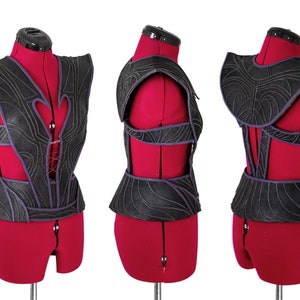 May include: A black leather corset with a purple trim and intricate detailing. The corset is on a red dress form.