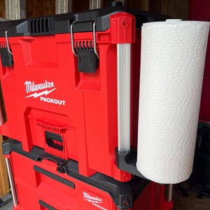 Peut inclure: Boîte à outils rouge Milwaukee Packout avec un porte-serviettes en papier blanc fixé sur le côté. Le porte-serviettes en papier est en métal et a une base en plastique noir.