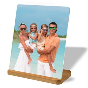 May include: A family of four, including two young girls, are standing on a beach. The father is holding one of the girls in his arms, while the mother is holding the other girl. All four are wearing sunglasses. The photo is displayed in a wooden frame.