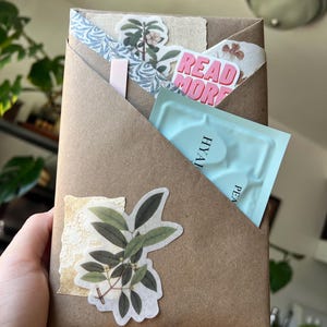 May include: A brown paper envelope decorated with botanical stickers, a piece of textured cream fabric, and a pink sticker that reads READ MORE. Inside the envelope is a light blue packet with text that includes HYAL and PEA.