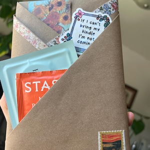 May include: A brown paper envelope containing a light blue packet, an orange tea bag, and a sticker with the text If I can't bring my kindle I'm not coming. The envelope is decorated with a sunflower print, floral tape, and a stamp of The Scream.