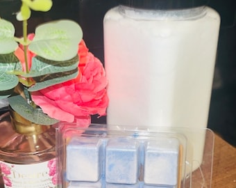 May include: A clear plastic container with six blue and white wax melts. The wax melts are in a grid pattern. The container is sitting on a wooden surface next to a white jar with a black lid.