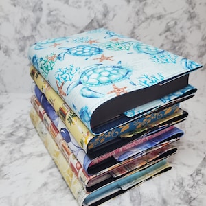 May include: A pile of book covers with diverse patterns. The top cover shows sea turtles, coral, and starfish on a light blue background. Other covers display floral, travel, and abstract designs. Each cover has a black interior and a fabric exterior.