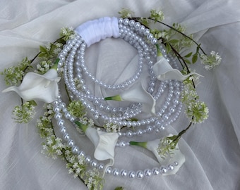 May include: A bridal bouquet featuring strands of faux pearls, white calla lilies, and small white flowers. The bouquet's handle is wrapped in white fabric. The arrangement is set against a white, textured background.