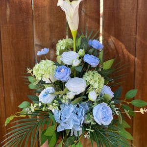 May include: A floral arrangement with a prominent white calla lily, light blue roses, and hydrangeas. The bouquet is complemented by green foliage. The flowers are arranged in a vase, creating a decorative display.