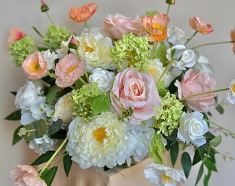 May include: A lush floral arrangement featuring a variety of flowers in shades of pink, peach, white, and yellow. The arrangement includes roses, peonies, and other blossoms, with green foliage. The flowers are arranged on a beige surface.