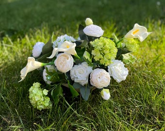May include: A white flower arrangement featuring calla lilies, roses, and hydrangeas. The flowers are arranged in a green vase and are sitting on a bed of green grass.
