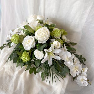 May include: A luxurious floral display composed of white roses, lilies, and orchids, complemented by green leaves and small green flowers. The arrangement is presented on a white surface, offering a delicate, refined aesthetic.