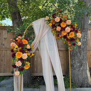 May include: A golden arch adorned with autumnal flowers in shades of orange, yellow, and burgundy. Soft, beige fabric drapes from the arch, forming a backdrop for an outdoor event. The arrangement is set against a wooden fence and trees, creating a natural setting.