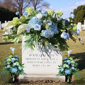 May include: A white headstone in a cemetery is decorated with a large arrangement of blue and white flowers. Two smaller floral arrangements are either side of the headstone. The headstone has engraved text.