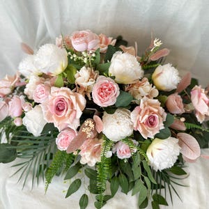 May include: A floral arrangement with pink roses, white peonies, and green foliage. The arrangement features various shades of pink and white flowers, with accents of green leaves and feathery pink accents. The flowers are arranged in a dense, natural style.