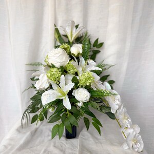 May include: A floral display with white lilies, roses, and orchids, complemented by green leaves. The flowers are arranged in a dark blue vase, creating a classic and elegant display. The arrangement is set against a white backdrop.