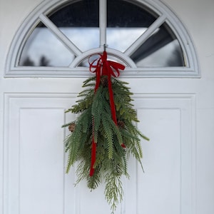 May include: A festive Christmas door decoration. A bundle of green pine and fir branches with pinecones, tied with a bright red ribbon, hangs from a white door with an arched window.