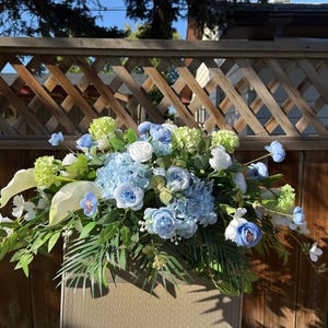 May include: A floral arrangement with blue, white, and green flowers. The arrangement includes hydrangeas, roses, and calla lilies, with green foliage. The flowers are arranged on a neutral-toned surface.