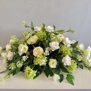 May include: A floral arrangement featuring white roses, light green hydrangeas, and delicate white blossoms. The flowers are arranged with dark green leaves, creating a natural and elegant display. The arrangement is set on a white surface.