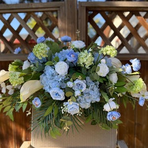 May include: A floral arrangement with blue hydrangeas, white roses, and green accents. The display includes white calla lilies and green foliage. The flowers are set against a wooden background. The arrangement is a mix of textures and colours.