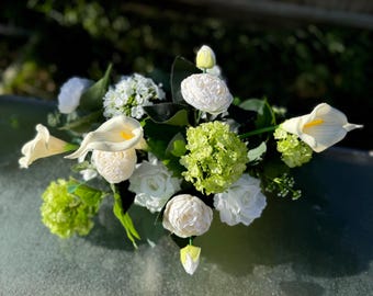 May include: A white flower arrangement featuring calla lilies, roses, and hydrangeas. The flowers are arranged in a vase and are surrounded by green foliage.