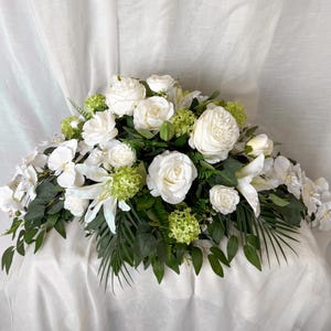 May include: A large floral arrangement with white roses, lilies, and other white flowers, complemented by green foliage and small green clusters. The arrangement is set against a white backdrop.