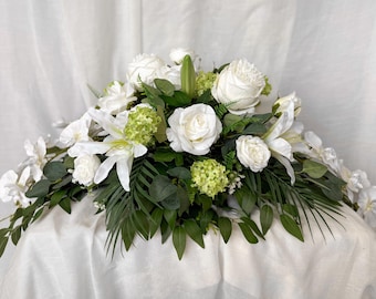 May include: A display of white roses, lilies, and other white flowers, complemented by green leaves. The arrangement is presented on a white surface, set against a white backdrop, offering a clean and elegant appearance.