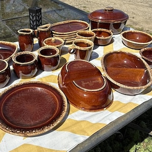 May include: A collection of brown ceramic serving dishes and bowls with a speckled glaze and a cream-colored trim. The set includes plates, bowls, mugs, and a covered casserole dish, arranged on a yellow and white striped tablecloth.