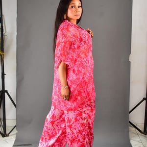 K&ouml;nnte beinhalten: Ein lebendiges rot-rosa gemustertes Ensemble, bestehend aus einem flie&szlig;enden Oberteil mit kurzen &Auml;rmeln und einer Hose mit weitem Bein. Das Outfit ist aus einem leichten Stoff gefertigt, ideal f&uuml;r warmes Wetter. Das Model tr&auml;gt goldene Ohrringe und ein Armband.