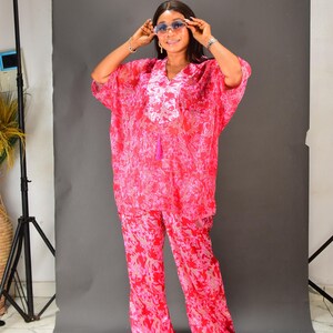 K&ouml;nnte beinhalten: Ein zweiteiliges Outfit mit rosa Blumenmuster. Das Oberteil ist eine locker sitzende Tunika mit kurzen &Auml;rmeln, V-Ausschnitt und Quastendetail. Die passende Hose hat weite Beine. Das Outfit wird mit blau get&ouml;nten Sonnenbrillen getragen.