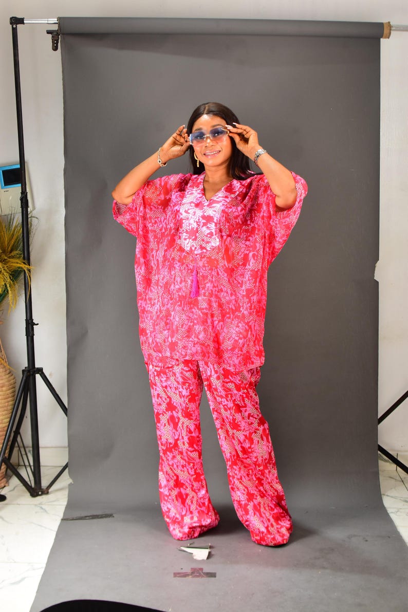 K&ouml;nnte beinhalten: Ein zweiteiliges Outfit mit rosa Blumenmuster. Das Oberteil ist eine locker sitzende Tunika mit kurzen &Auml;rmeln, V-Ausschnitt und Quastendetail. Die passende Hose hat weite Beine. Das Outfit wird mit blau get&ouml;nten Sonnenbrillen getragen.