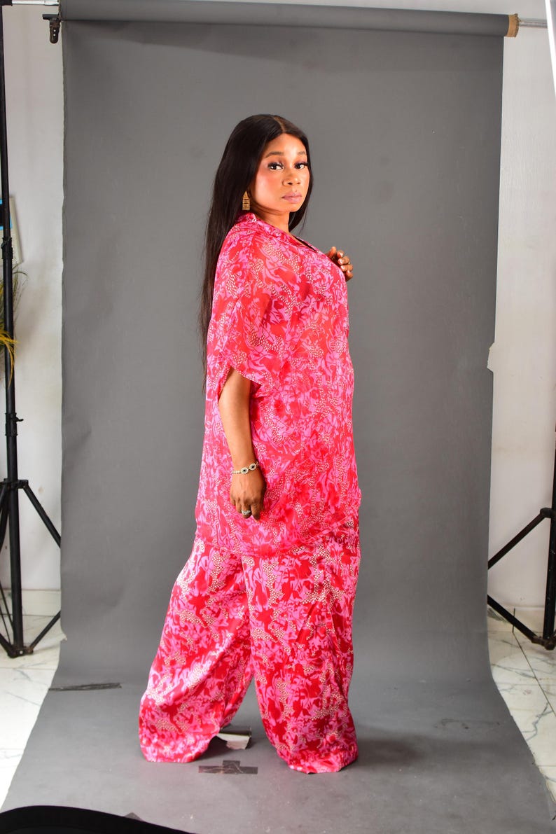 K&ouml;nnte beinhalten: Ein lebendiges rot-rosa gemustertes Ensemble, bestehend aus einem flie&szlig;enden Oberteil mit kurzen &Auml;rmeln und einer Hose mit weitem Bein. Das Outfit ist aus einem leichten Stoff gefertigt, ideal f&uuml;r warmes Wetter. Das Model tr&auml;gt goldene Ohrringe und ein Armband.