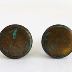 May include: Two circular cabinet knobs with a weathered, dark bronze finish. The surface of each knob displays a mottled texture with visible patches of oxidized teal green patina along the edges and across the flat, round faces of the hardware.