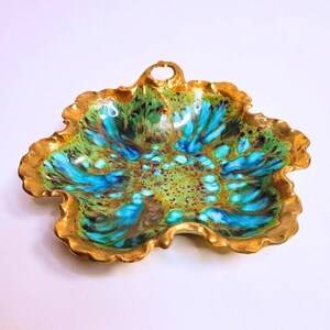May include: A decorative ceramic dish with a scalloped, ruffled edge finished in a metallic gold glaze. The interior features a vibrant, mottled glaze pattern in shades of bright turquoise, deep forest green, and brown, with a small handle loop.