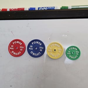 Könnte beinhalten: Vier farbige Gewichtsscheiben in Rot, Blau, Gelb und Grün, jeweils mit dem Wort "POWER" und dem Gewicht in Kilogramm. Die Scheiben sind in absteigender Reihenfolge des Gewichts angeordnet: 25, 20, 15 und 10 kg. Das Wort "LIFTING" ist ebenfalls sichtbar.