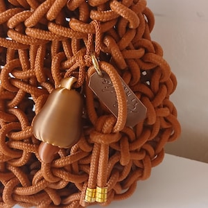 May include: A brown handbag crafted from thick, knotted rope with a dark brown handle. The bag features a decorative charm shaped like a pear with a metallic finish, a small rectangular brown tag, and two gold-toned metal beads on a cord.