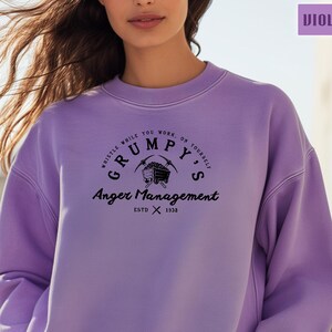 May include: A purple sweatshirt with a black graphic design that says "Whistle while you work on yourself. Grumpy's Anger Management. Est. 1938." The graphic features a cartoon image of a mine cart and pickaxe.