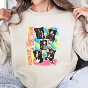 May include: A white sweatshirt with a graphic print of the band New Kids on the Block. The print features a collage of images of the band members in a colorful, abstract design. The text "New Kids on the Block" is printed in pink and black.