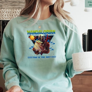 May include: A light blue sweatshirt with a graphic of the Mandalorian and Grogu. The graphic is in a triangle shape with the text "Mandalorian" at the top and "This is the way" at the bottom.