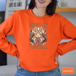 May include: Orange sweatshirt with a graphic design of two crossed guitars with flowers and the text "Nashville Tennessee".