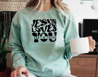 May include: A light blue sweatshirt with the text "Jesus Loves You And I'm Trying" in black block letters.
