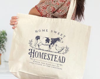 May include: Cream-colored tote bag with a black and white illustration of a cow and chicken, with the words "HOME SWEET HOMESTEAD" and "WHERE THE KIDS ARE AS FREE RANGE AS THE CHICKENS" printed on it. The bag has a simple design and a long strap.