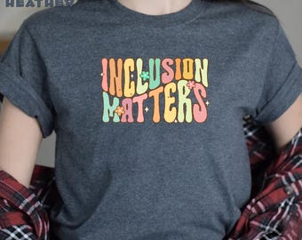 May include: A dark heather grey t-shirt with the text "INCLUSION MATTERS" in a retro, colorful font. The text is decorated with small flowers and sparkles.