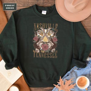 May include: A dark green sweatshirt with a graphic print of two guitars crossed over each other with flowers and the text "Nashville Tennessee".