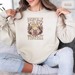 May include: A cream colored sweatshirt with the text "Nashville Tennessee" and a graphic of two crossed guitars with flowers and a sun.